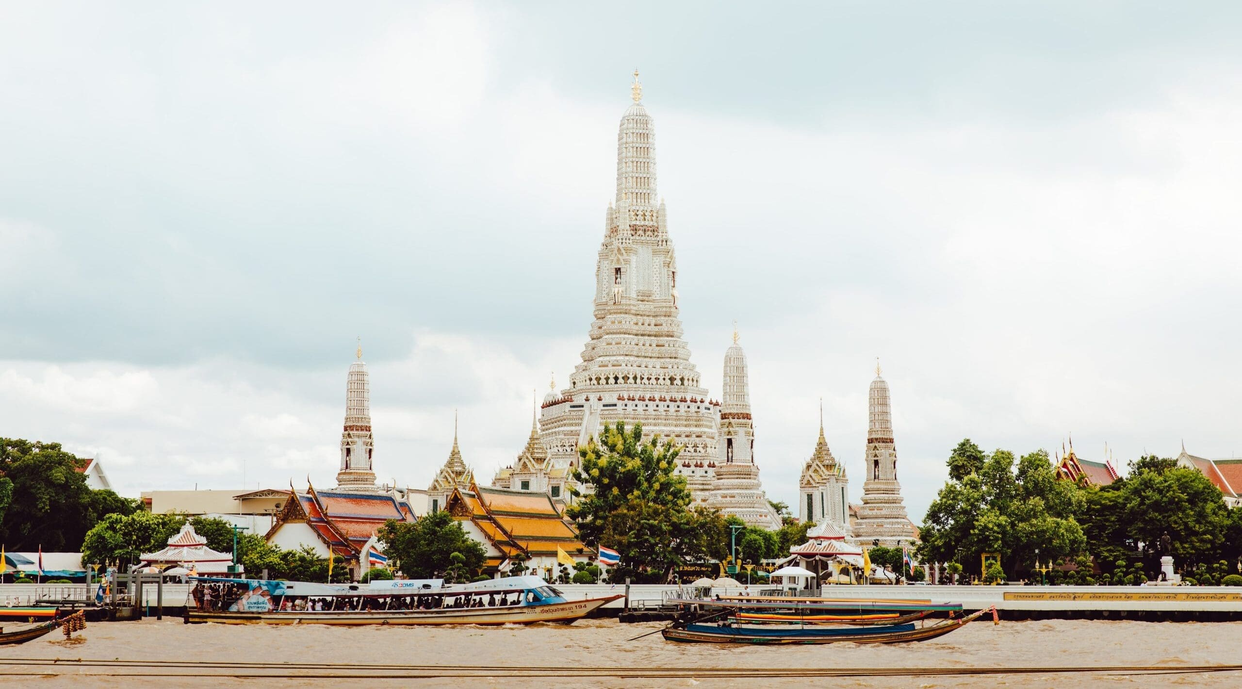 Wat Arun, a Buddhist temple in Bangkok, features a central spire surrounded by smaller towers, with trees and roofs in the foreground and cloudy sky above